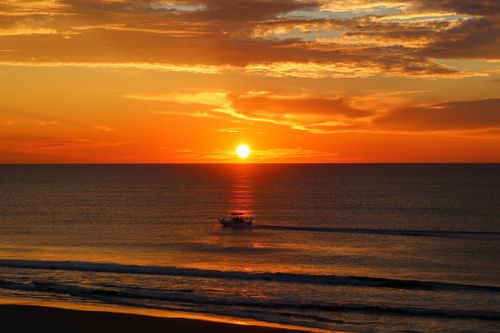 Free A stunning sunset over the ocean with a silhouetted boat on the horizon and vibrant sky reflections. Stock Photo