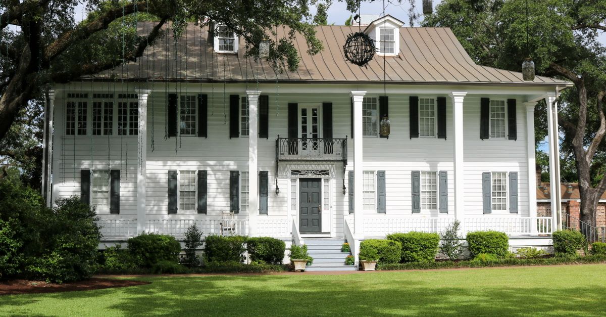 Elegant colonial house with lush lawn and classic facade surrounded by trees.