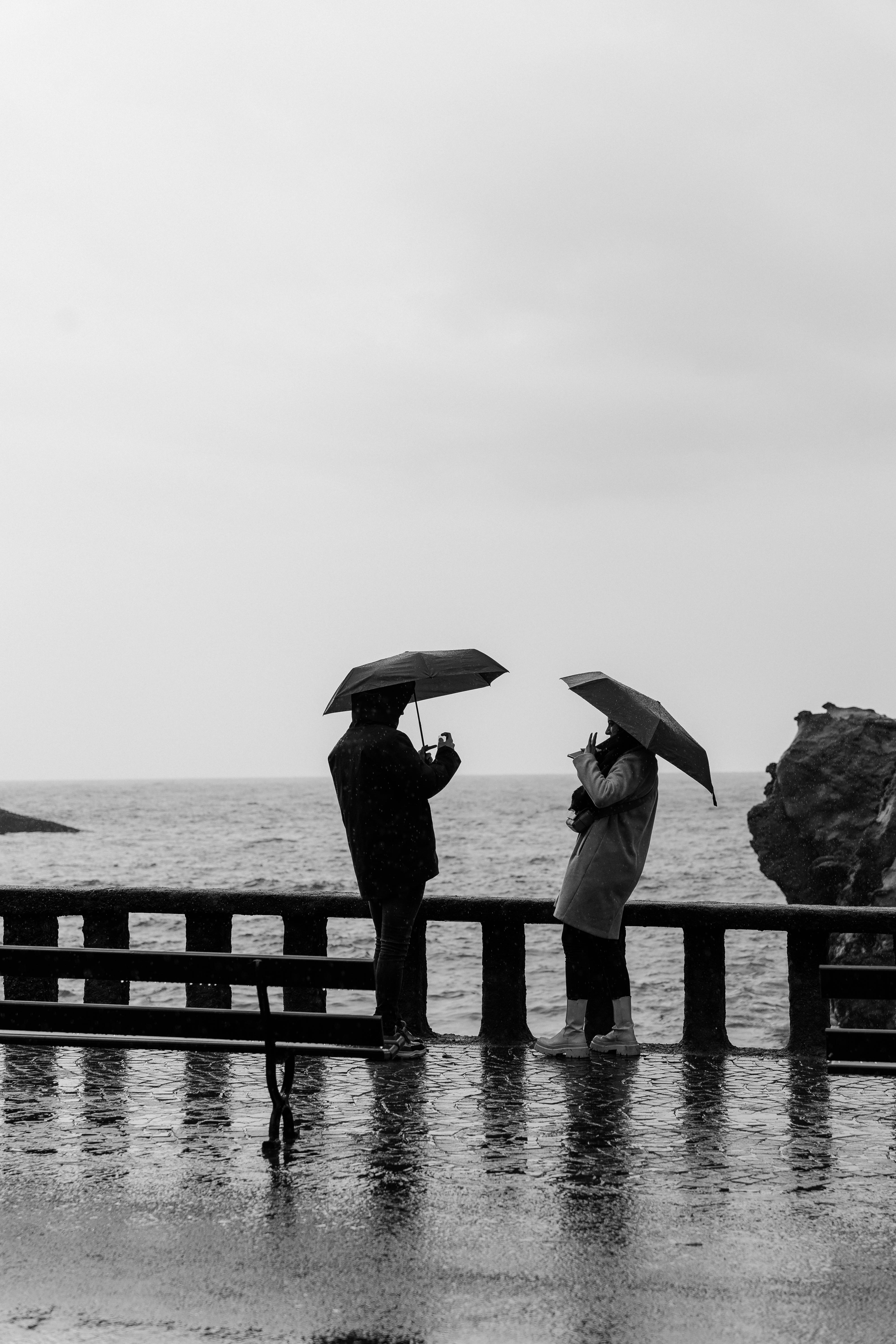 Monochrome photo of a couple with umbrellas by the ocean, capturing a rainy, serene moment.