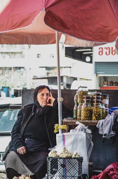 A woman selling pickles at a street market stall, shaded by a pink umbrella.