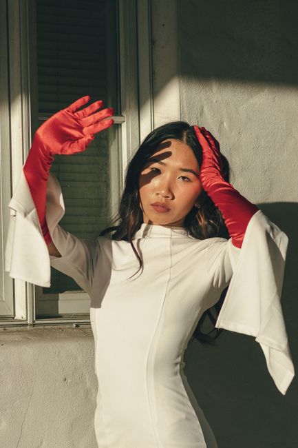 Portrait of an Asian woman in a white dress and red gloves posing stylishly in natural light.
