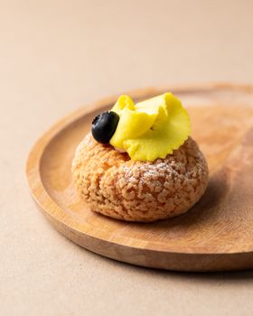Elegant cream puff topped with yellow frosting and a blueberry on a wooden tray.
