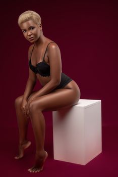 Striking studio shot of a model in black lingerie posed on a stool against a rich red backdrop.