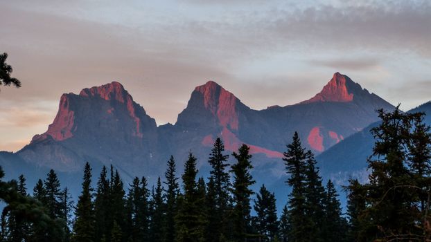 Beautiful sunrise over the Three Sisters peaks in Canmore, Alberta, with a forest silhouette.