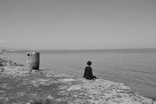A lone figure sits on a cliff edge, gazing over the serene ocean horizon in a monochrome setting.