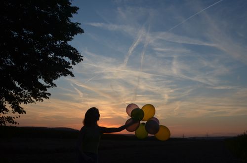 Free A person holding colorful balloons silhouetted against a vibrant sunset sky. Stock Photo