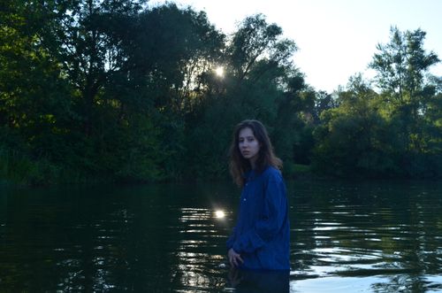 Free A woman stands in a tranquil river surrounded by lush trees during sunset, creating a peaceful atmosphere. Stock Photo