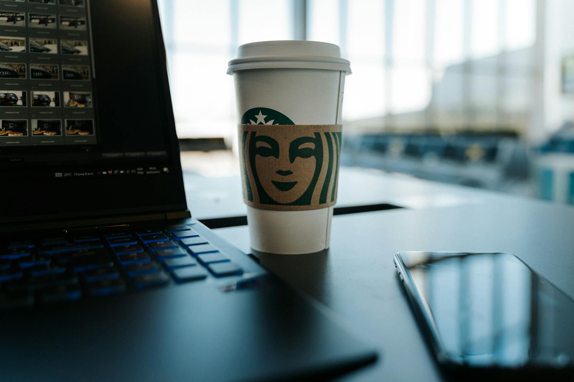 A disposable coffee cup next to a laptop and phone in a modern airport setting.