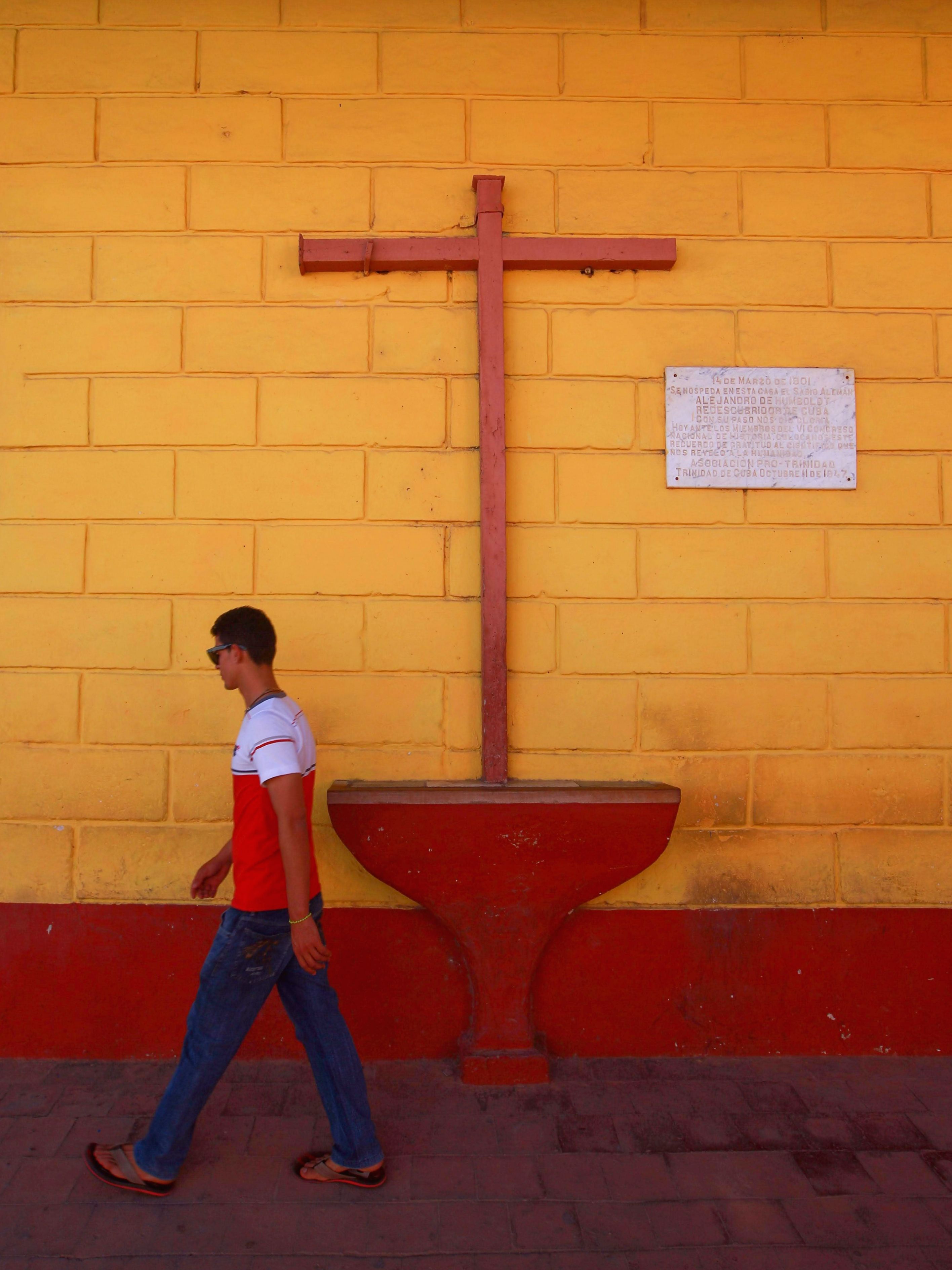 A man walks by a vibrant yellow wall with a prominent wooden cross, creating a mix of color and symbolism.