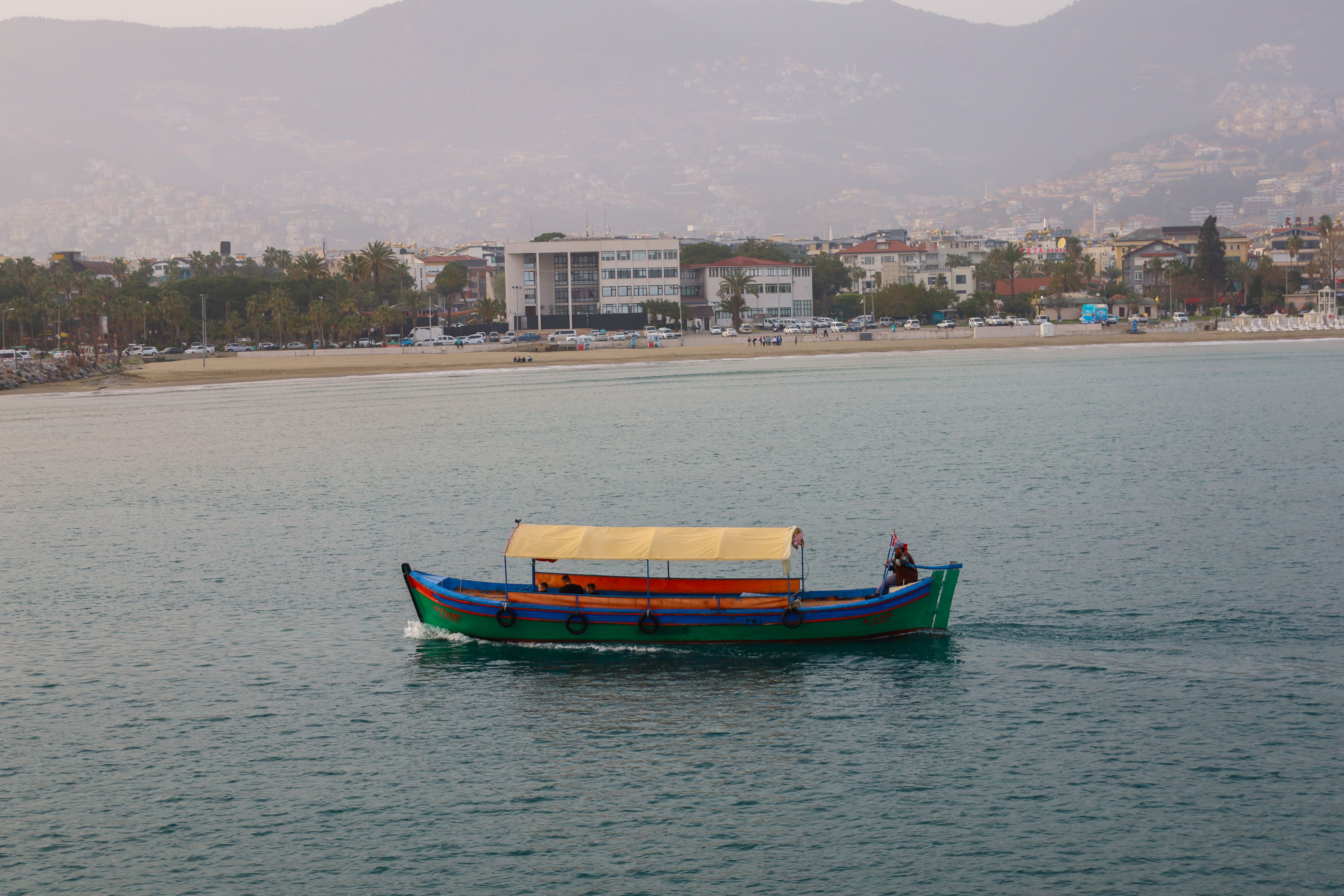 A colorful traditional boat sails on Antalya's coastal waters, with the cityscape in the background.