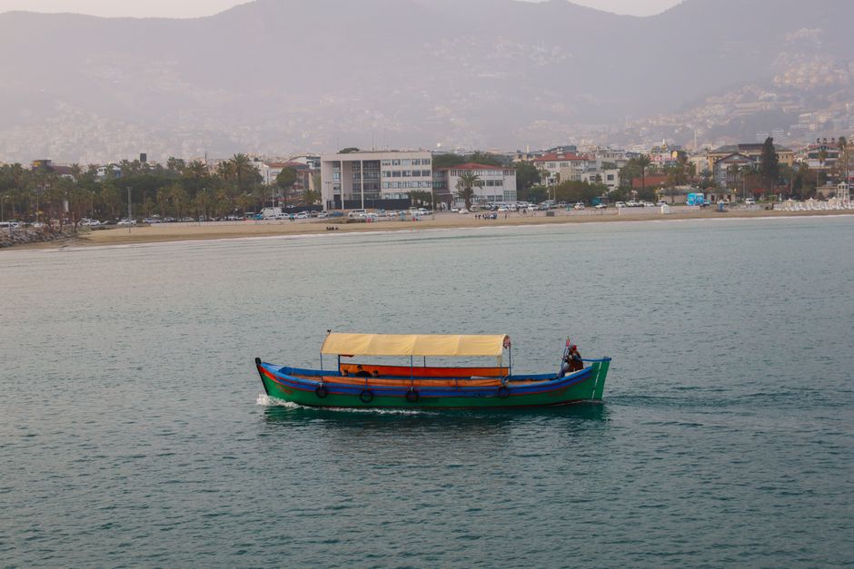 A colorful traditional boat sails on Antalya's coastal waters, with the cityscape in the background.