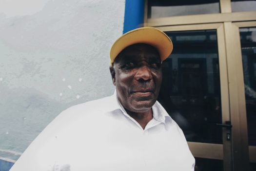 Close-up of an elderly man wearing a yellow cap and white shirt, standing outdoors.