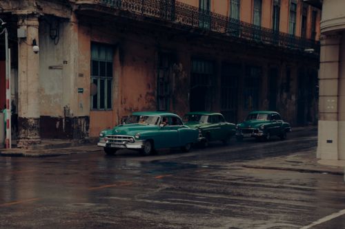 Free Classic green cars parked alongside a shabby, historic building on a wet urban street. Stock Photo