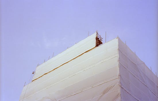 A building under renovation wrapped in protective material with visible scaffolding against a clear sky.