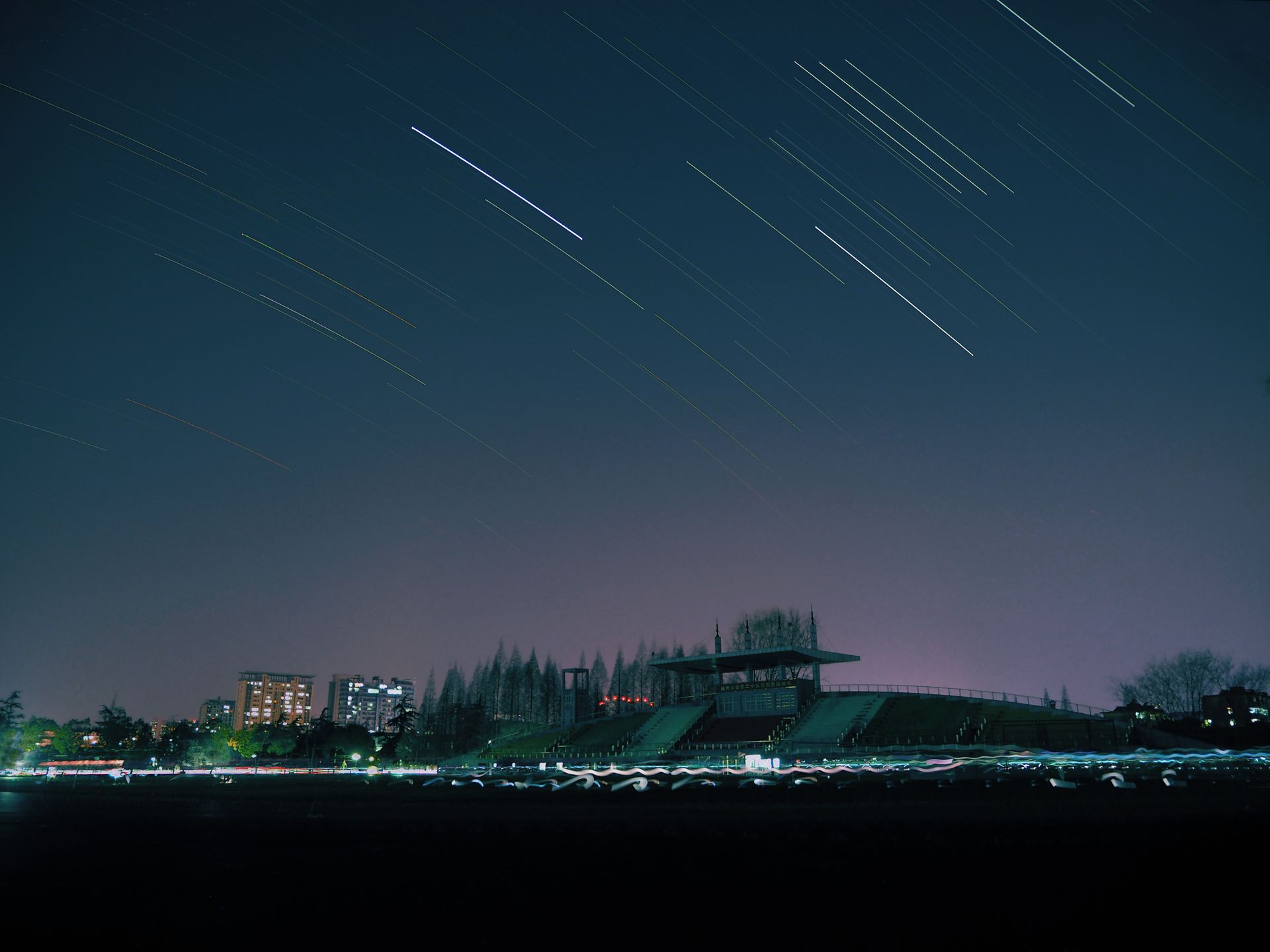 A breathtaking view of a stadium under a starry night sky with impressive star trails and city lights.