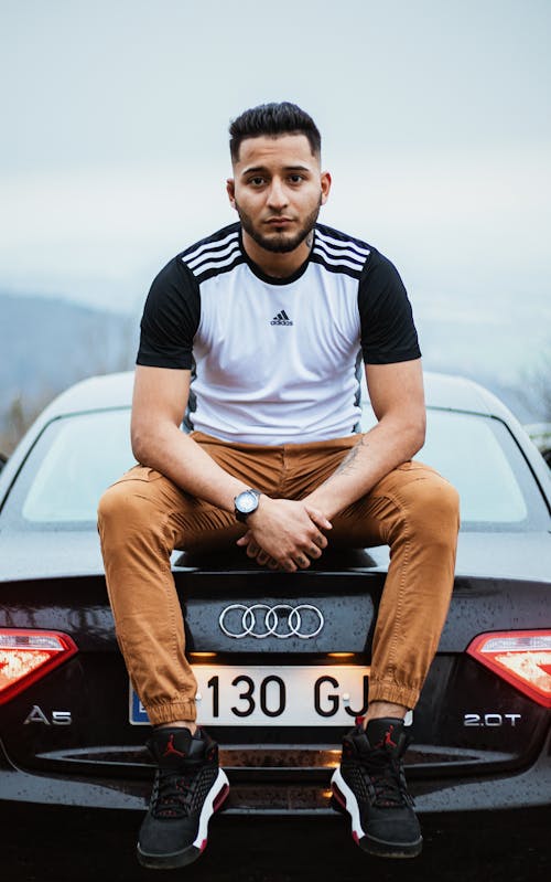 Young Man in Adidas T-shirt and Brown Pants Sitting on Car Trunk