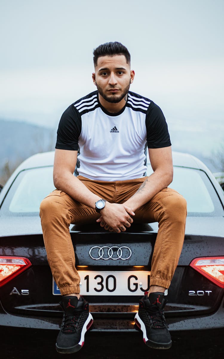 Young Man In Adidas T-shirt And Brown Pants Sitting On Car Trunk