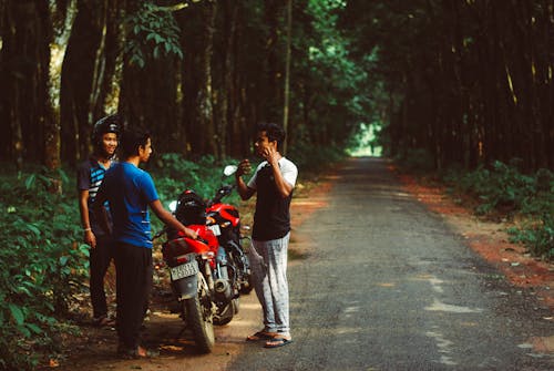 三名男子站在紅色的摩托車旁邊，綠樹環繞
