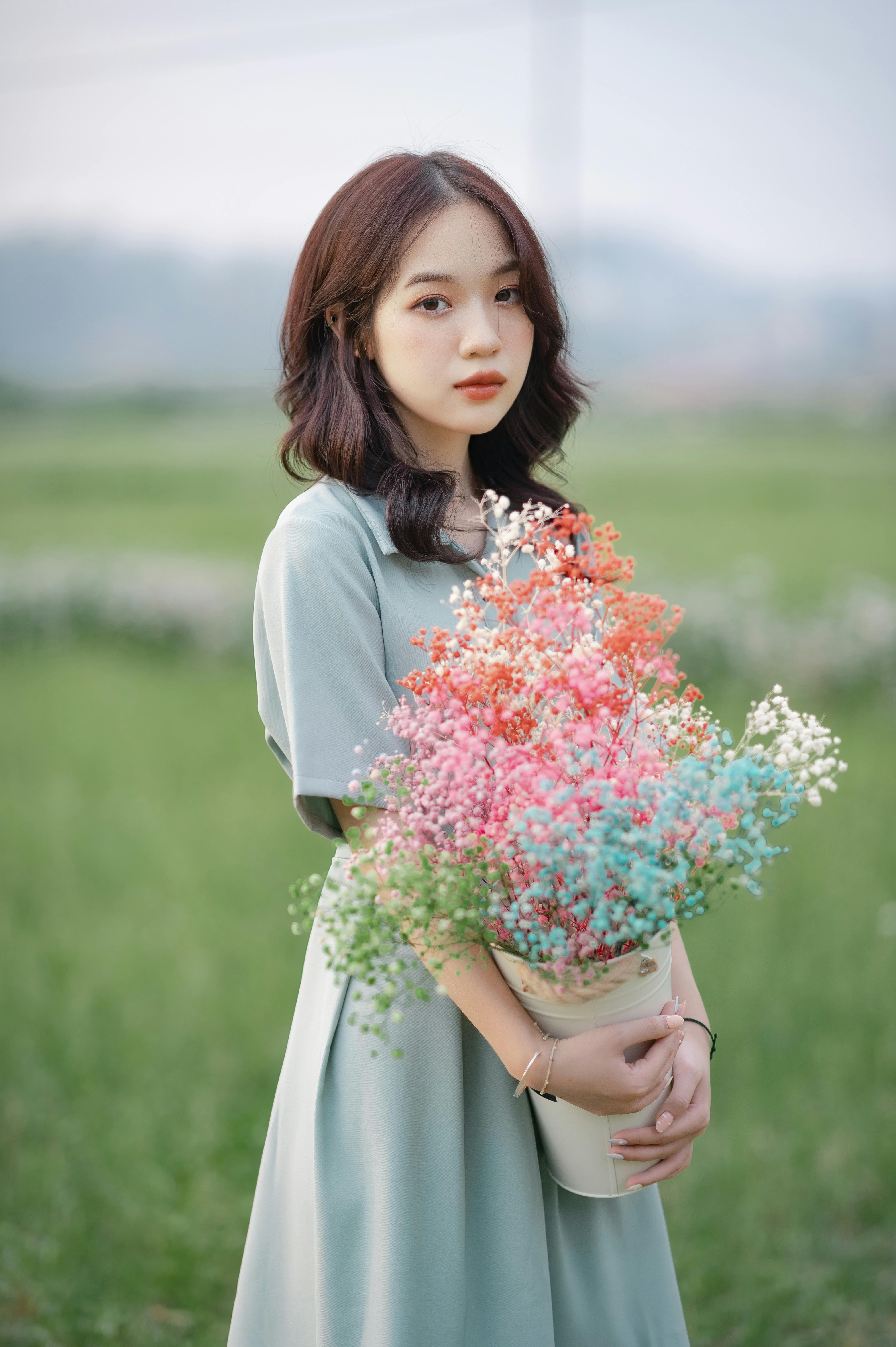 Asian woman in a green dress holding a bouquet of colorful flowers in a lush outdoor setting.