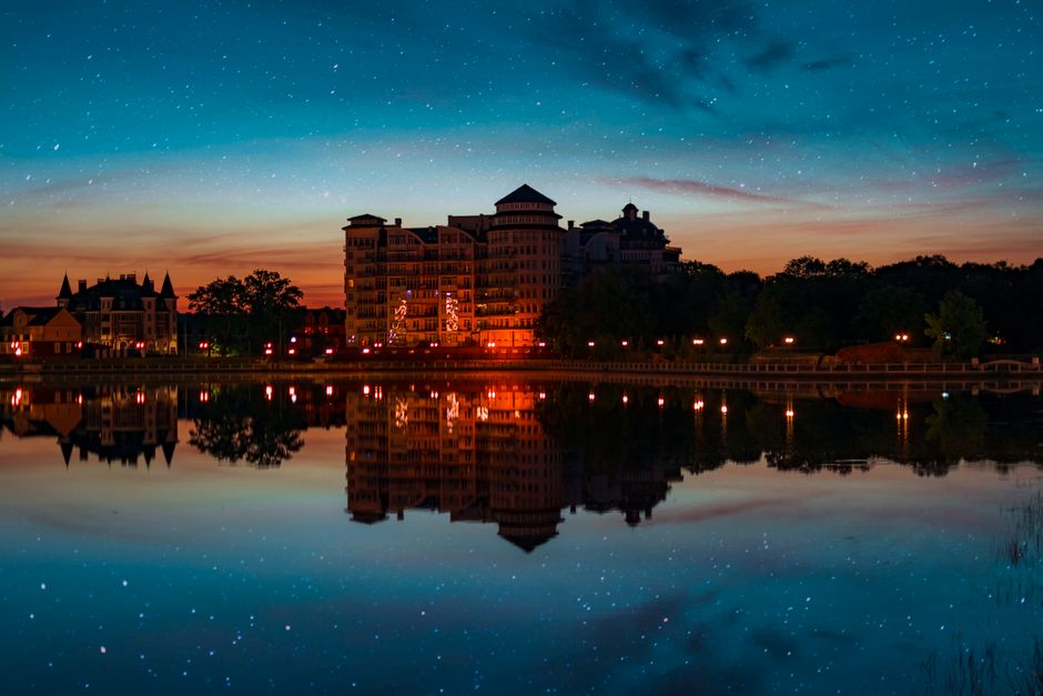 Illuminated buildings reflecting on calm water during a starry twilight sky.