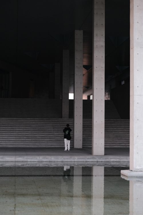 Free A solitary figure stands in modern architecture with a reflective pool, creating a moody ambiance. Stock Photo