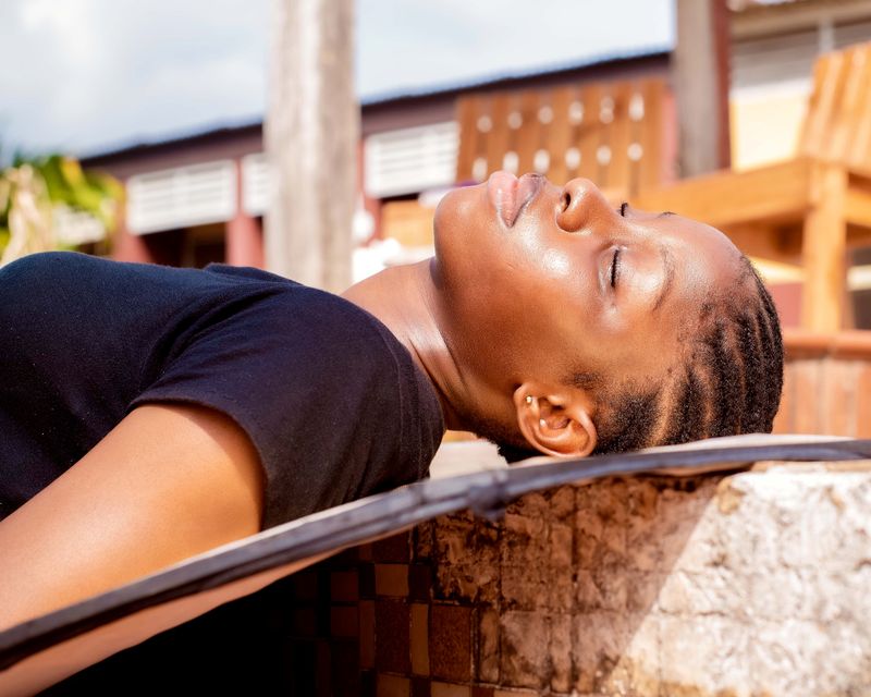 A Black woman with braided hair rests her head back against a low wall in bright sunlight, eyes closed, wearing a dark T-shirt, her neck and face shining with sweat on a hot day.
