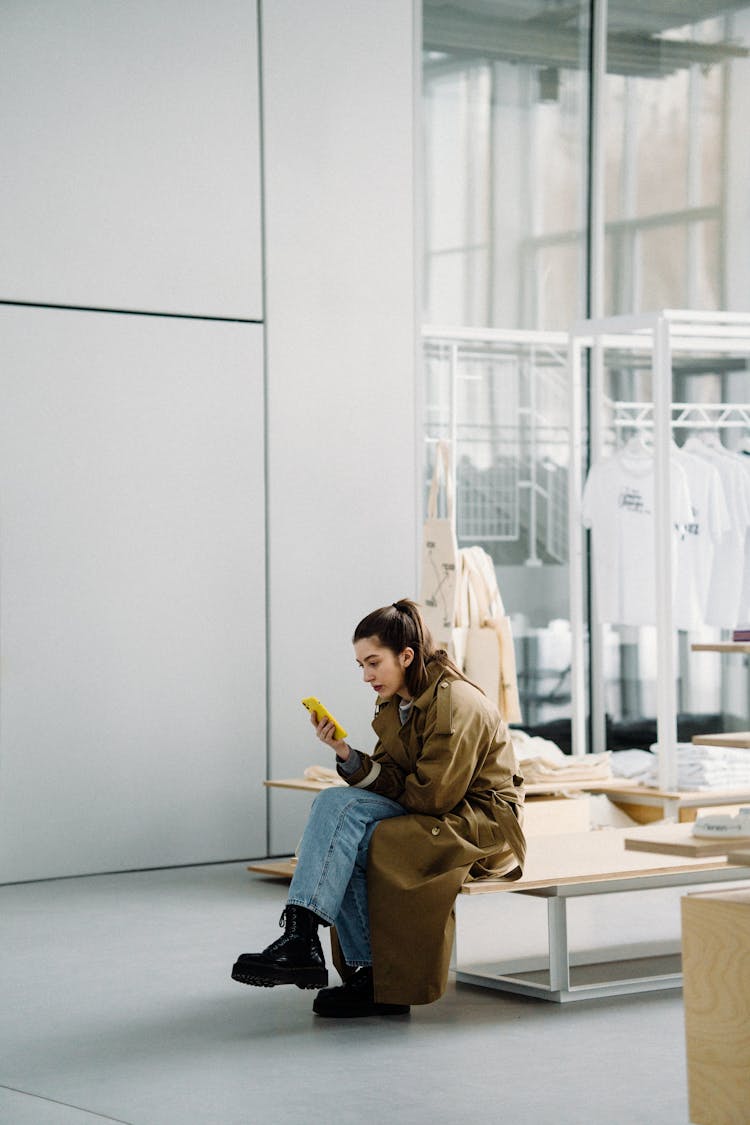 Woman In Coat Sitting Alone On Bench And Texting On Mobile Phone