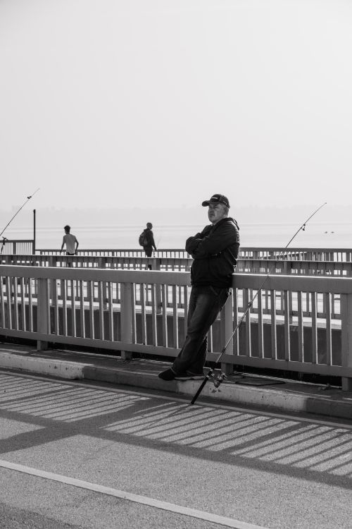 Free Black and white image of a man leaning on a railing while fishing from a bridge. Stock Photo