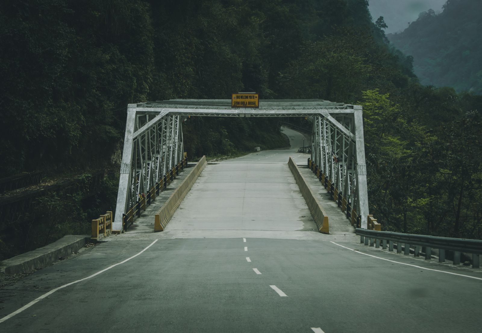 A steel truss bridge carrying an empty road across a river gorge in the forested hills of Sikkim