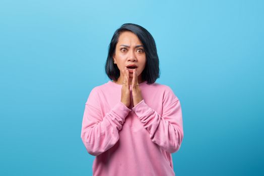 Surprised woman in a pink sweatshirt with hands on chin against a bright blue backdrop, expressing shock.