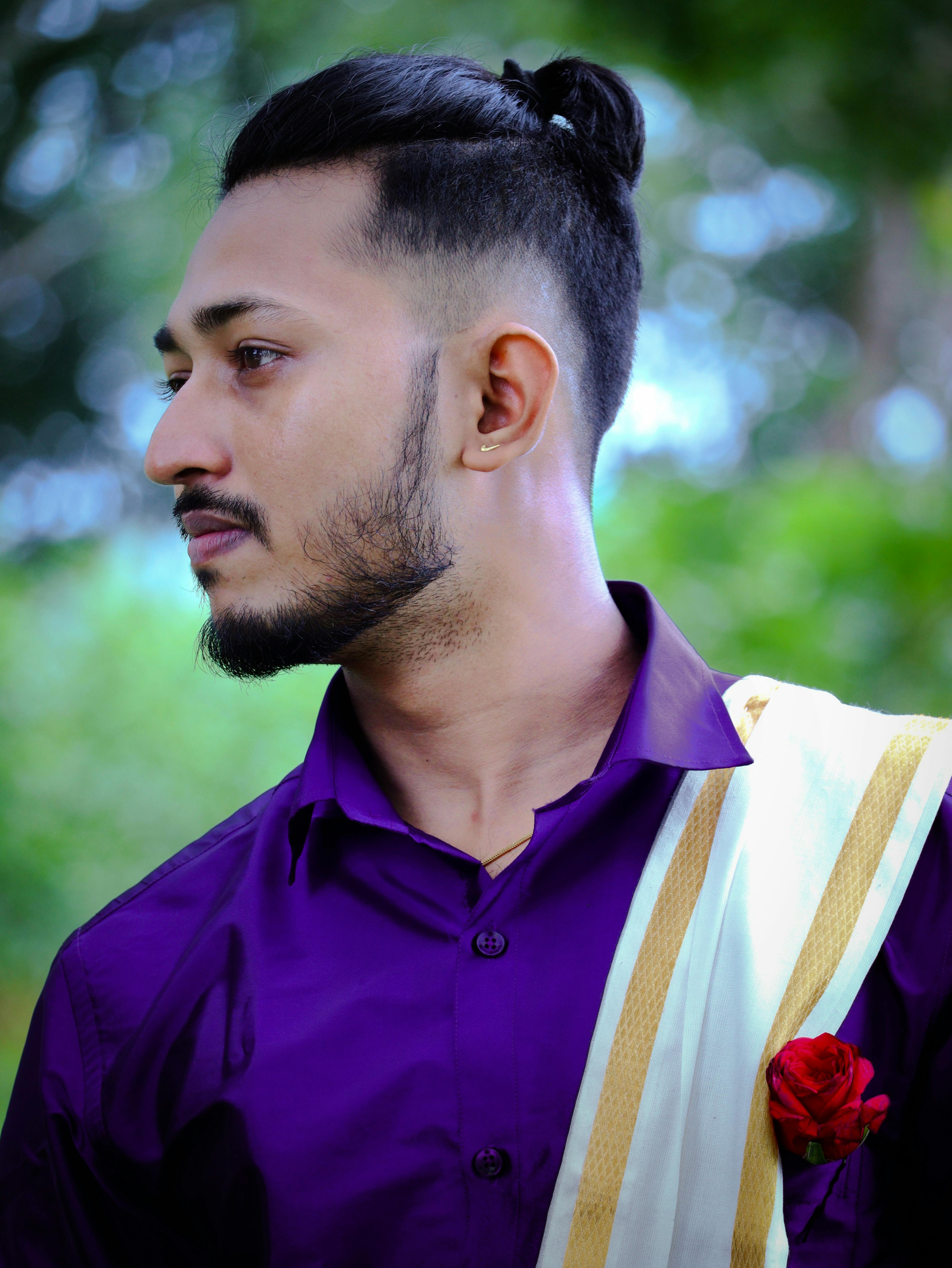Side profile of a stylish bearded man with a man bun wearing a purple shirt and sash, in an outdoor setting.
