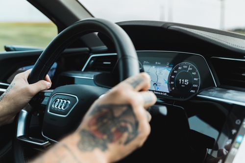 Free Close-up of a tattooed driver handling a car with an Audi steering wheel and digital display showing 115 km/h. Stock Photo