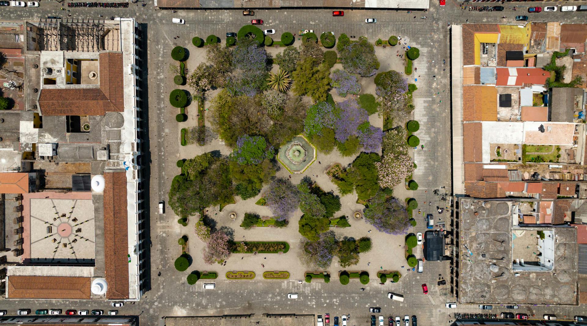 Drone shot of a vibrant city square with trees and well-designed gardens surrounded by urban architecture.