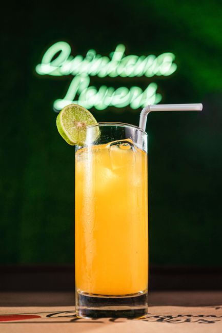 A tall glass of orange cocktail with ice and lime wedge against a green neon background.