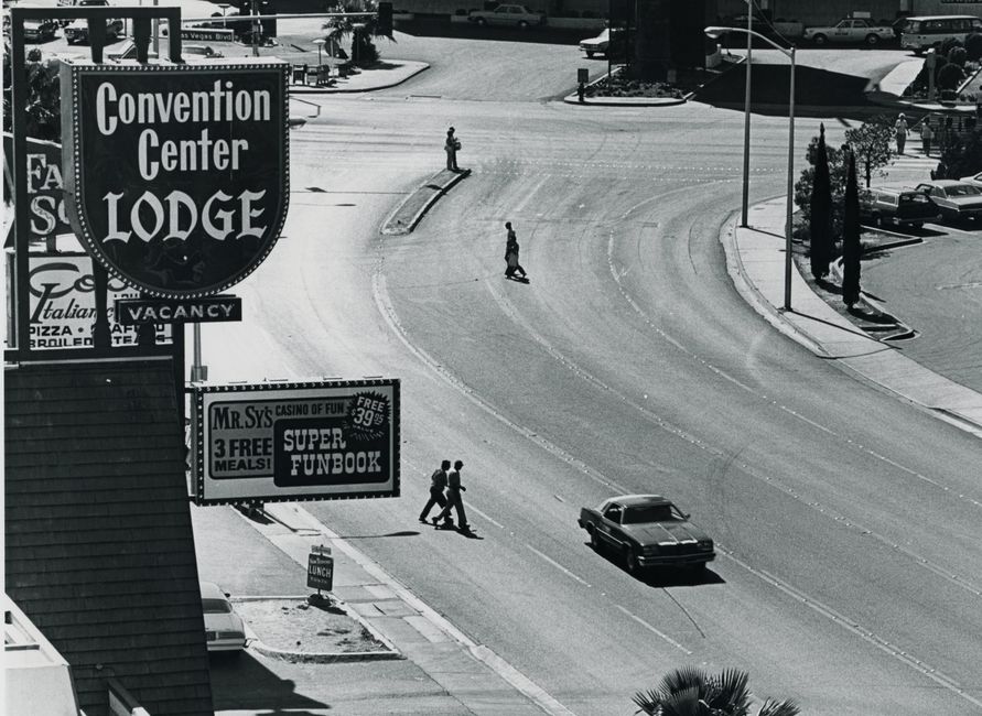 A black and white image capturing a vintage street scene in Las Vegas with pedestrians, cars, and prominent signage.