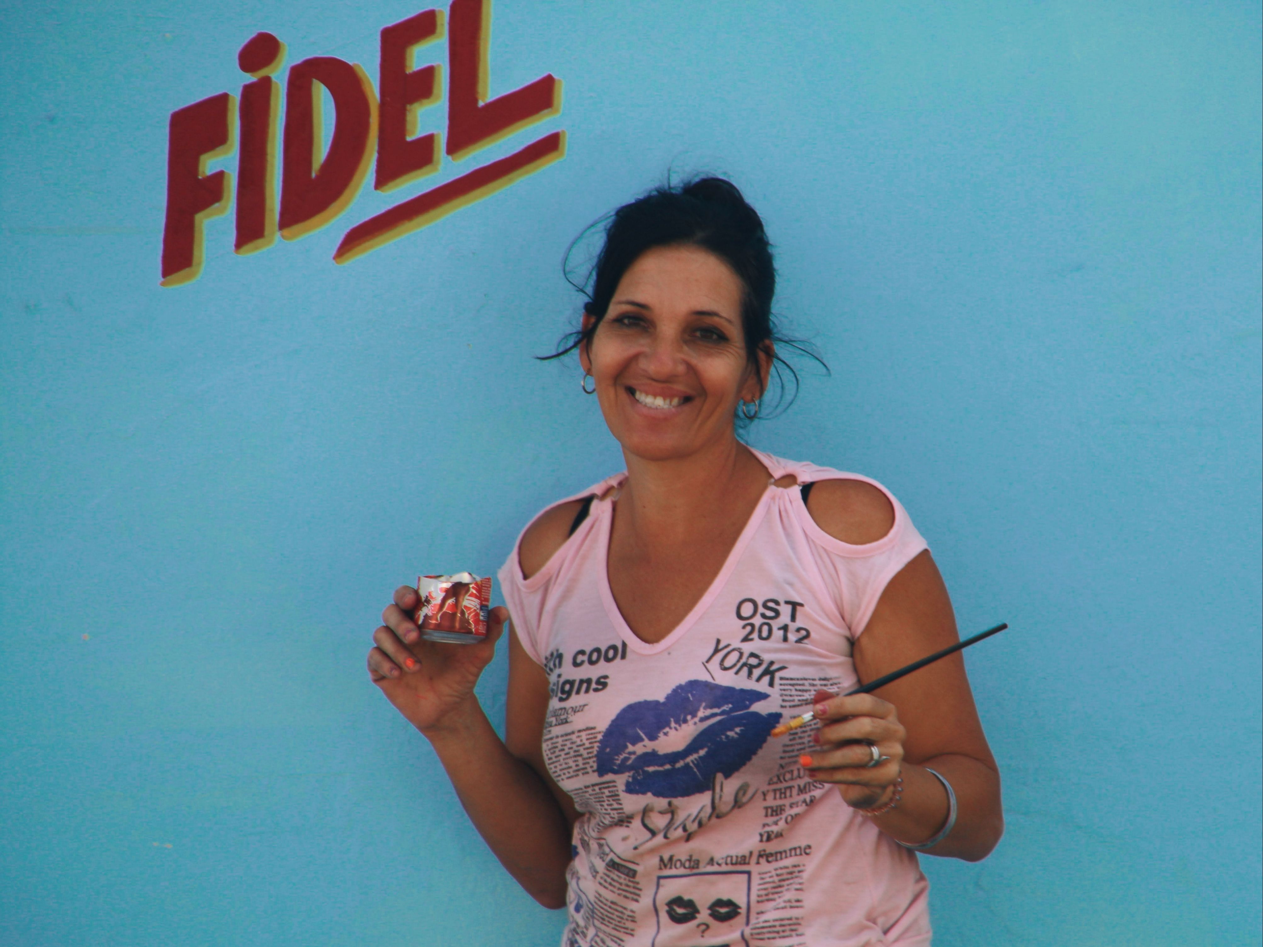 A cheerful woman stands against a blue wall, holding a paint can and brush, with 'Fidel' painted above.