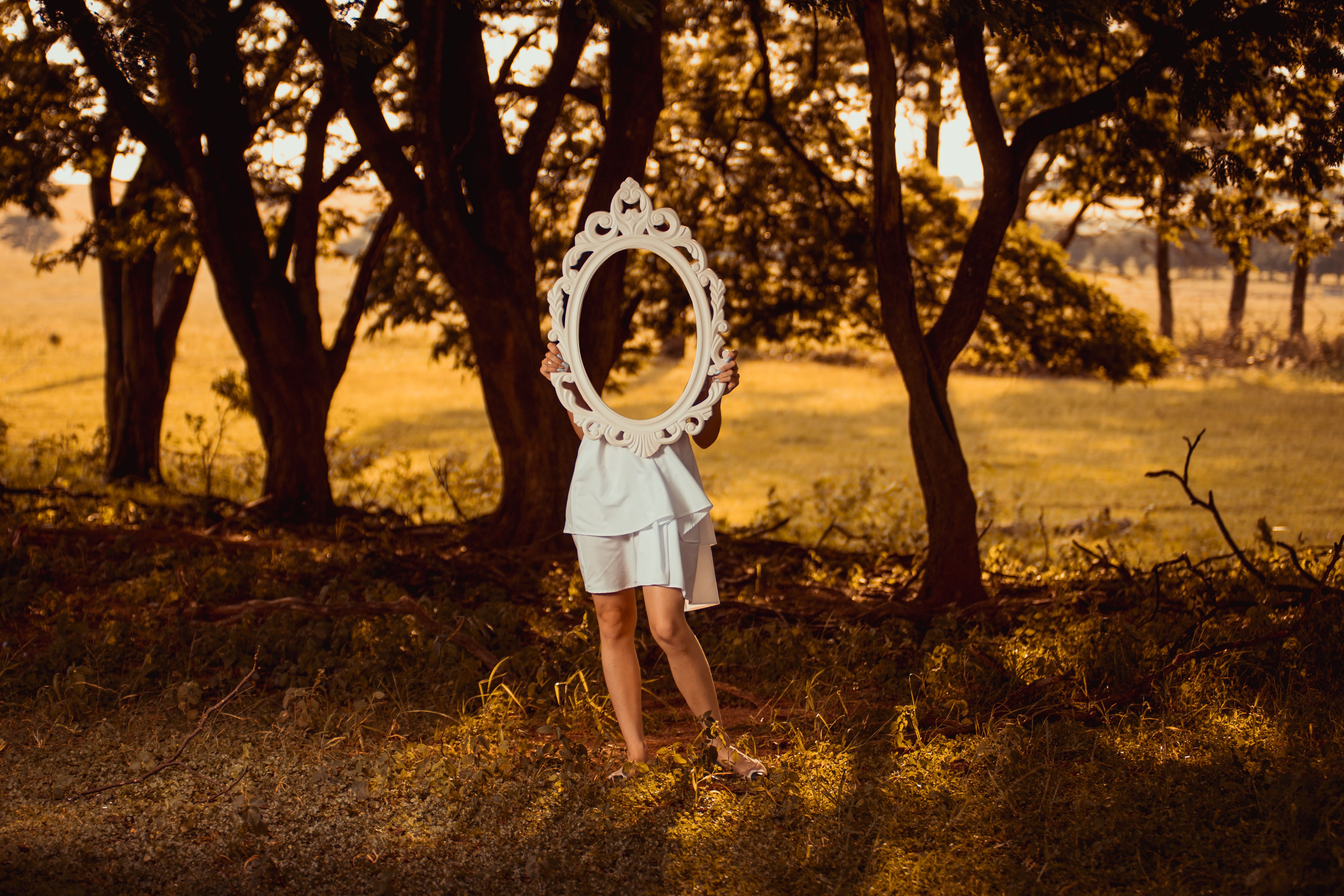 A mysterious woman holding an ornate mirror in a sunlit forest, creating a whimsical reflection.