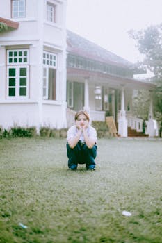 A teenager sits thoughtfully on grass in front of a bright white house.