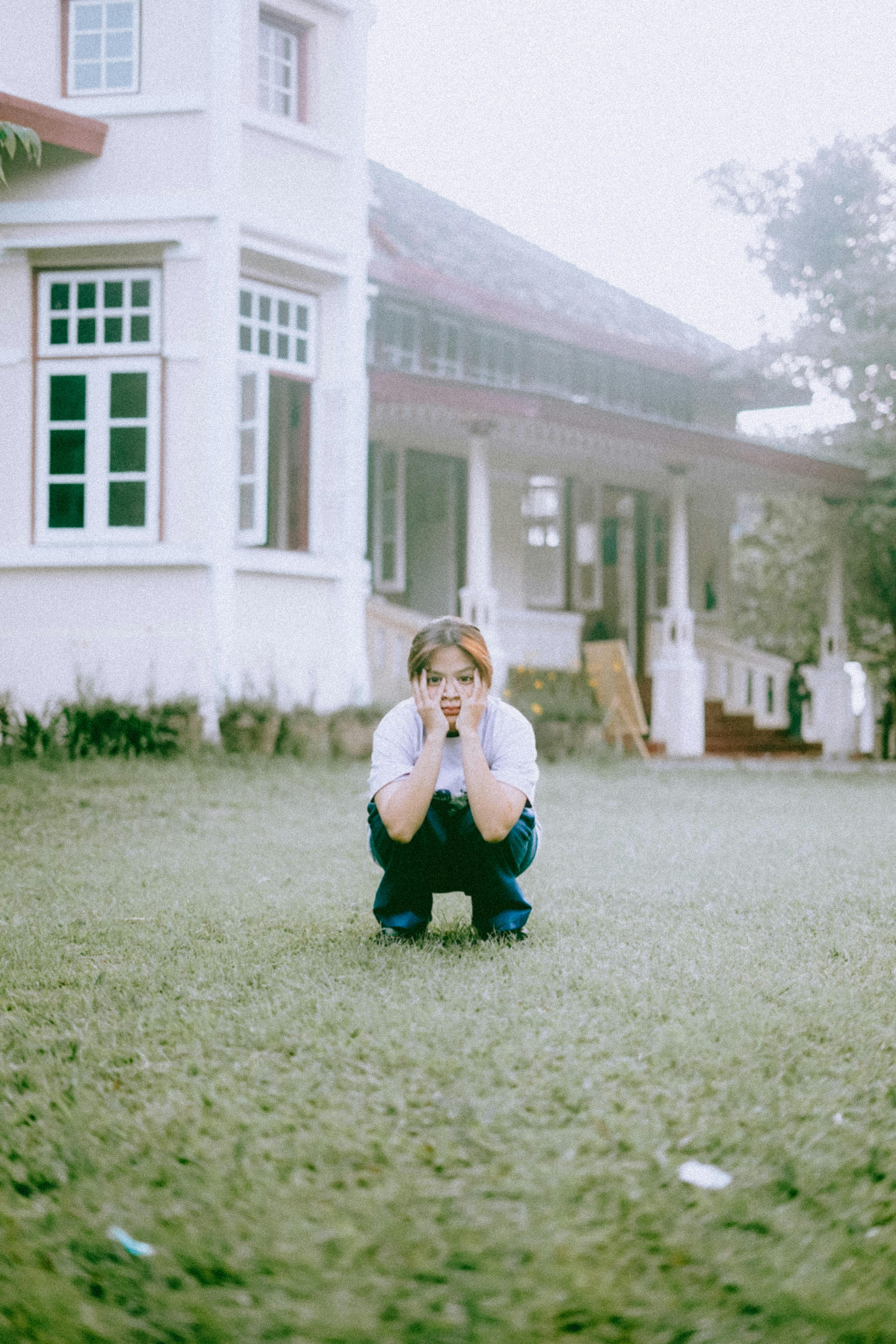 A young woman crouches on a grassy lawn, surrounded by a vintage house in an outdoor setting.