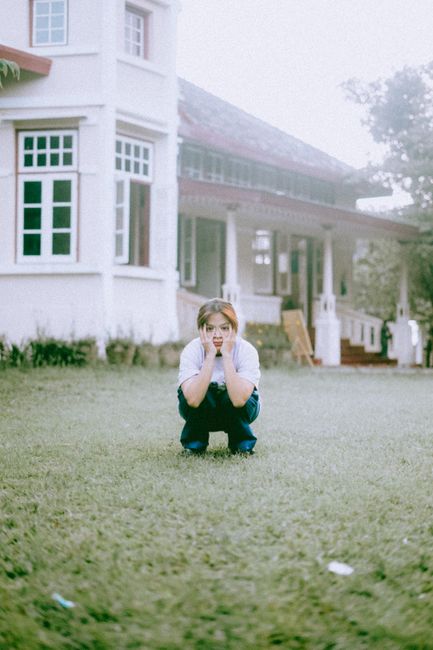 A young woman crouches on a grassy lawn, surrounded by a vintage house in an outdoor setting.