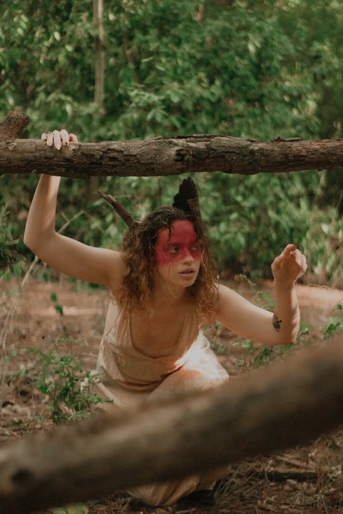 Woman in Face Paint Crouching in Forest Holding on to a Tree Branch