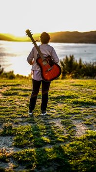 A woman stands with a guitar near a serene lake at sunset in Kawakawa Bay, New Zealand.
