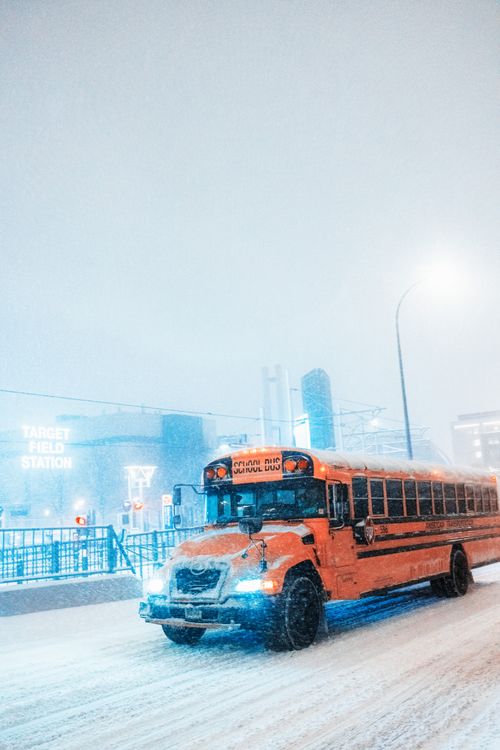 Free A school bus navigates a snow-covered road amidst city buildings during a heavy snowfall. Stock Photo