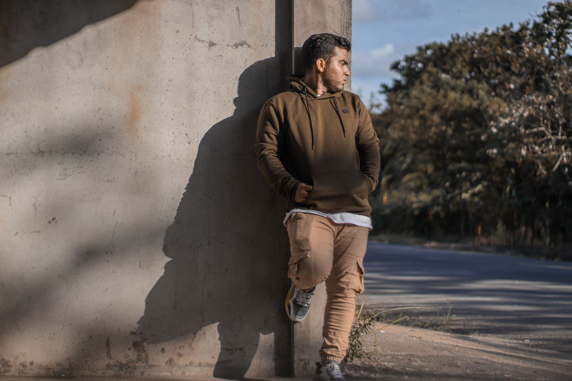 Homme en hoodie kaki et cargo beige adossé à un mur béton en tenue streetwear décontractée