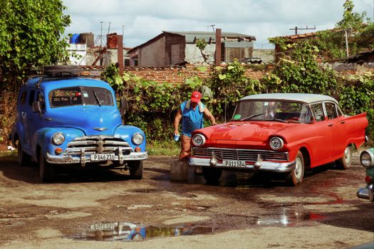 A man washing two vintage cars outdoors, with water reflecting the classic scene.
