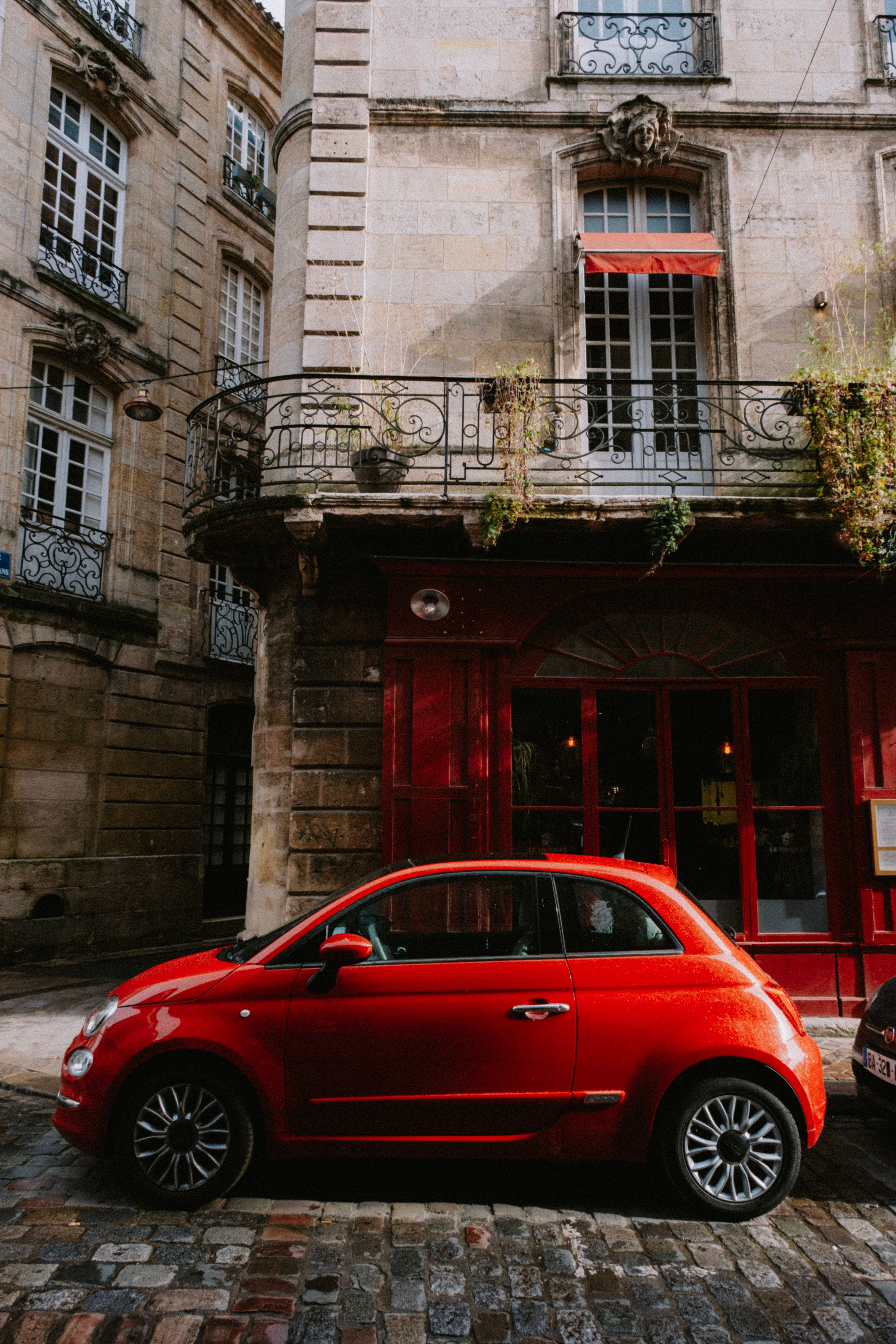 A vibrant red car parked beside a classic European building with baroque balconies.