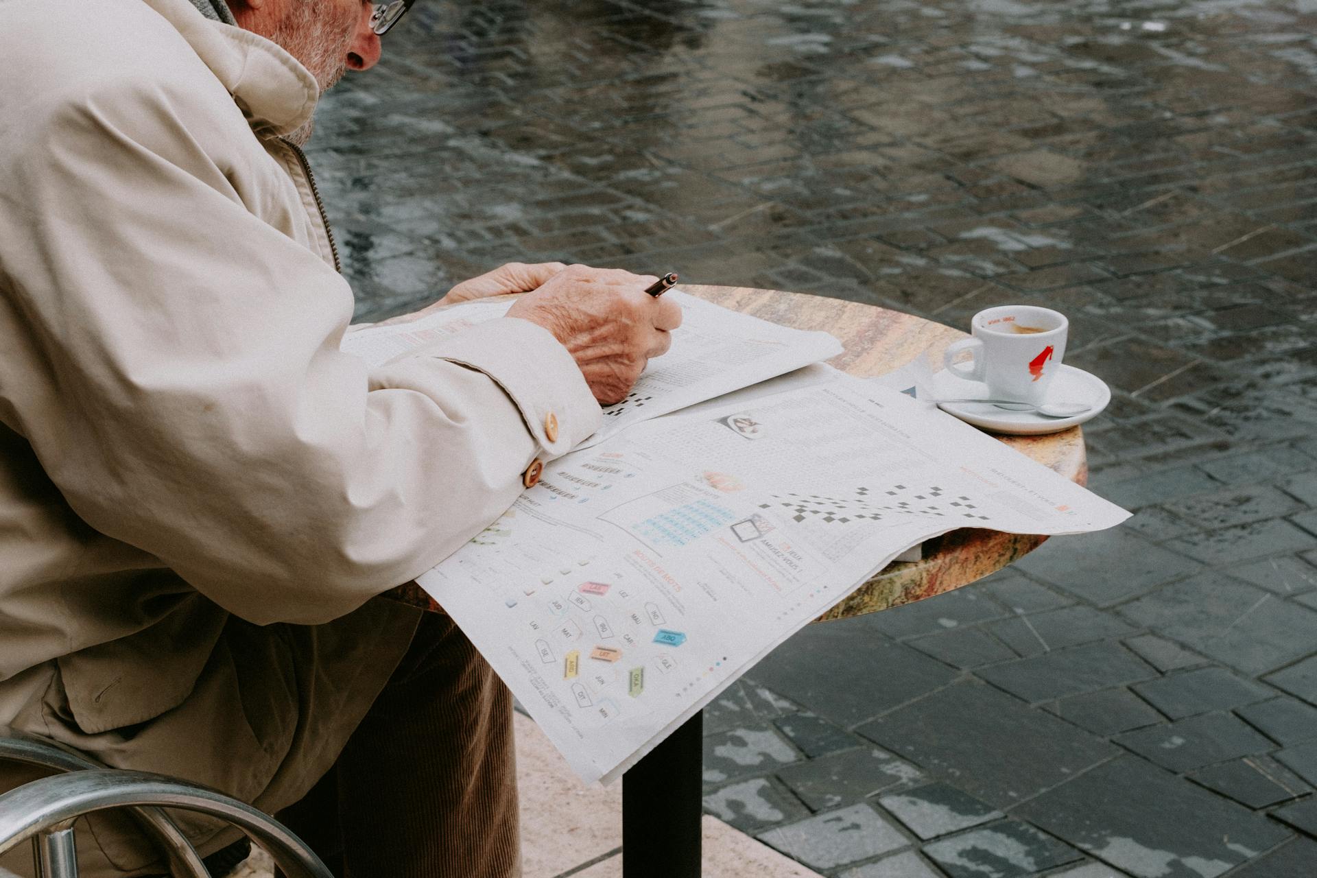 A senior man enjoys leisure time solving a crossword puzzle at an outdoor cafe.