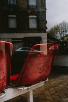 A moody urban scene with reflections of red chairs and street view through a window.