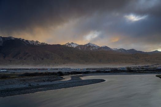 A stunning mountain range with snowcapped peaks and dramatic clouds at sunset, featuring a calm river in the foreground.