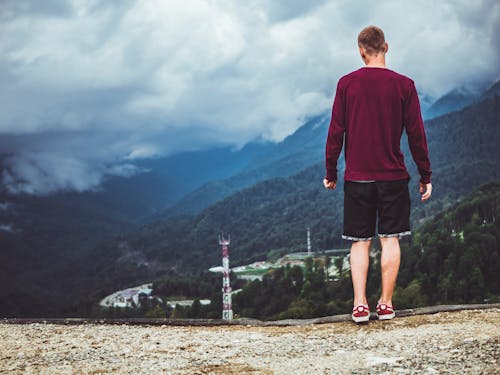 Pria Mengenakan Sweater Maroon Dan Celana Pendek Hitam Berdiri Di Depan Gunung